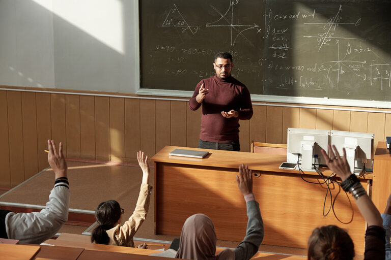 Middle aged teacher in casualwear standing by blackboard in lecture hall and looking at students